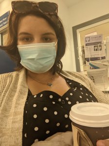 Picture of Rebecca wearing a facemask and holding a takeaway coffee cup sitting in a hospital waiting room waiting for an appointment. Wearing a brown cardigan, black white spotty top, blue disposal facemask, 