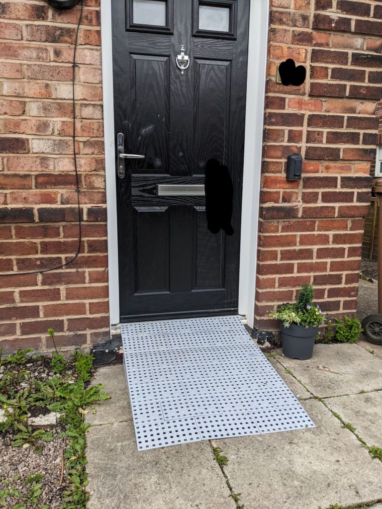 A black front door with a grey ramp leading up to it. The house number is scribbled out with black lines for privacy 